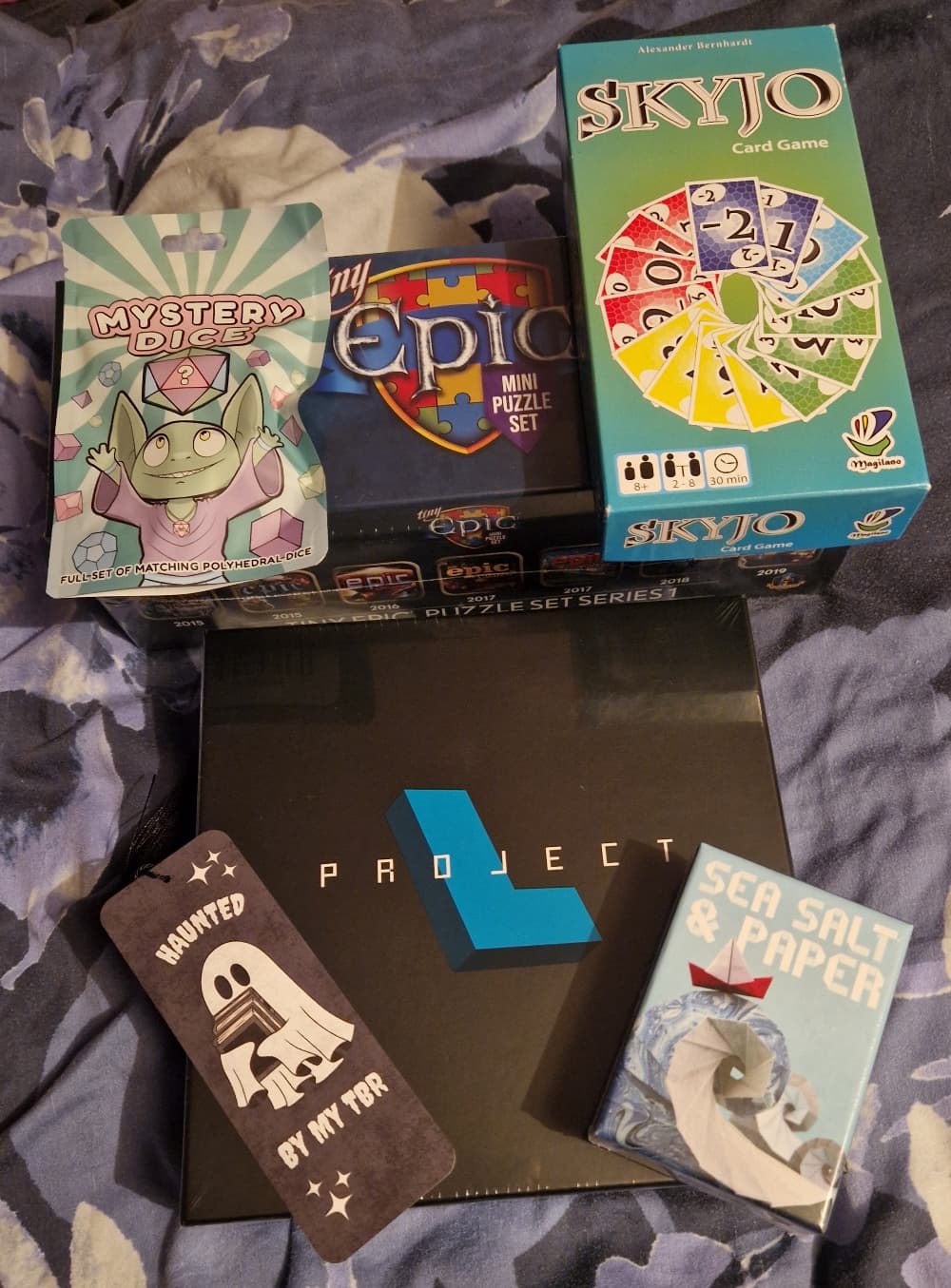 Games and other items arranged in the middle of a blue duvet: Project L, Sea Salt and Paper, Skyjo, a packet of dice, a box of Tiny Epic jigsaws, and a black bookmark with an illustration of a ghost